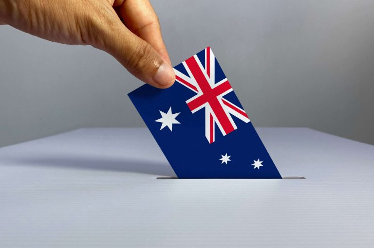 Voting / election concept: hand holding voting card with country national flag, inserting it into white ballot box; clean neutral background — Australia